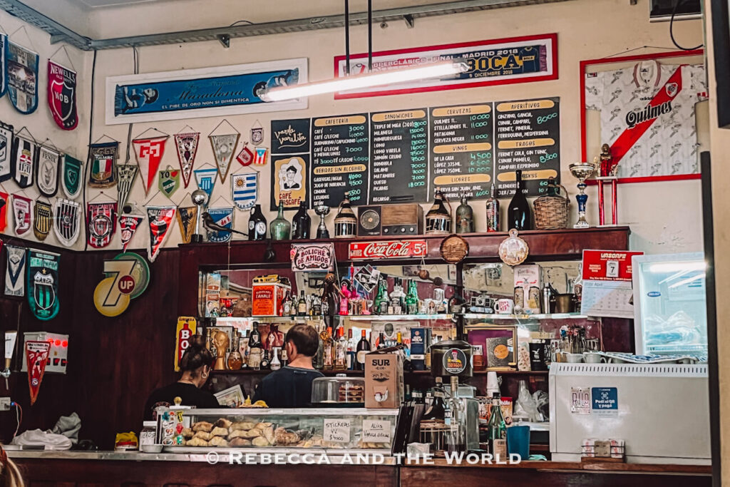 Interior of Bar Notable Varela Varelita in Buenos Aires, featuring walls covered with vintage football pennants, team banners, framed memorabilia, and old advertising signs. Behind the wooden bar counter, shelves are packed with bottles, trophies, photos, and retro decorations, while handwritten chalkboard menus display drinks and sandwiches overhead.