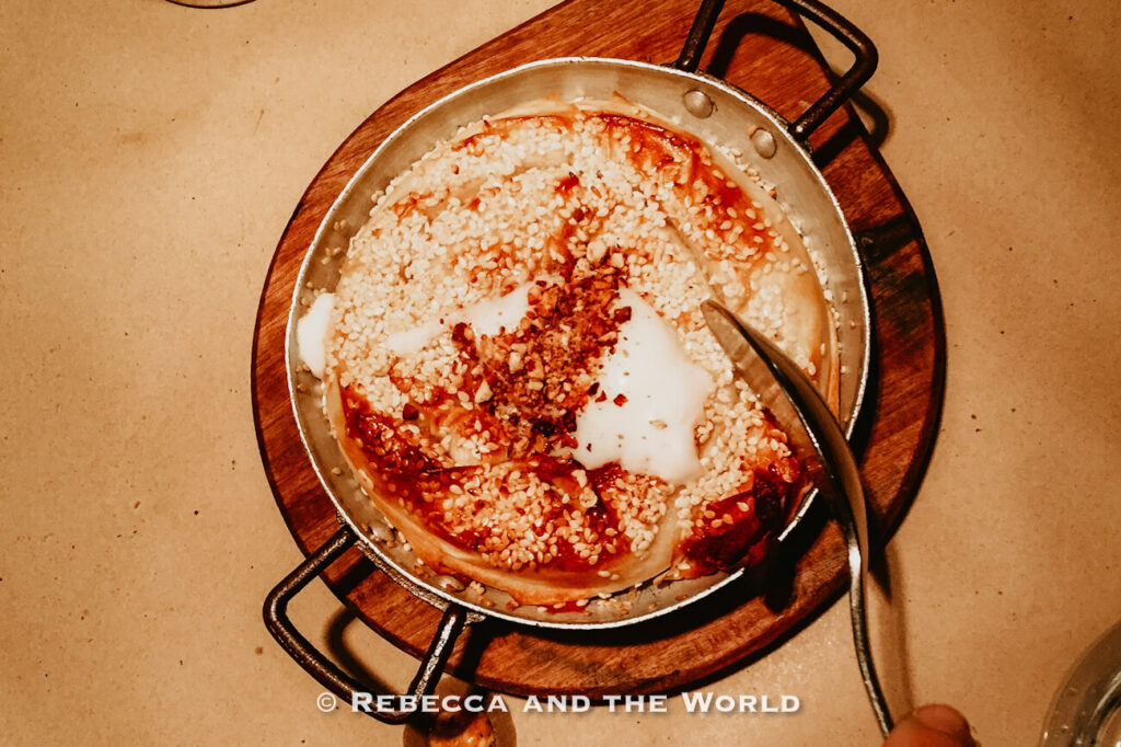 Overhead view of a pastry baked in a round metal pan set on a wooden serving board. A spoon rests inside the pan. The warm restaurant lighting highlights the rich textures at one of the best places to eat in Buenos Aires for creative Jewish food, Mishiguene.