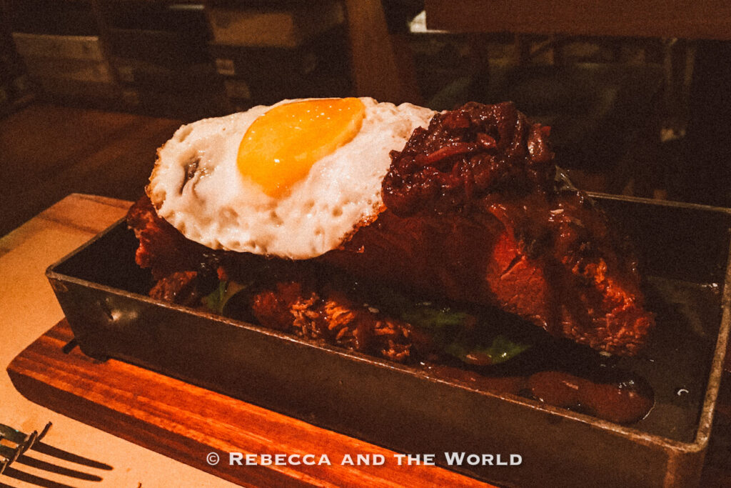 Close-up of a hearty pastrami dish served in a rectangular black tray, topped with a sunny-side-up fried egg and dark glossy sauce. The dramatic low lighting and rustic presentation showcase elevated comfort food at stylish Buenos Aires restaurants.