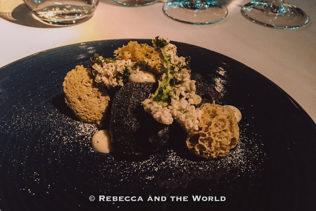 Gourmet plated dish served on a dark blue plate with black pasta or dumplings surrounded by airy crisps, creamy sauce, and leafy garnishes. Powdered seasoning is dusted across the plate while wine glasses sit blurred in the background, highlighting artistic presentation at some of the best places to eat in Buenos Aires.