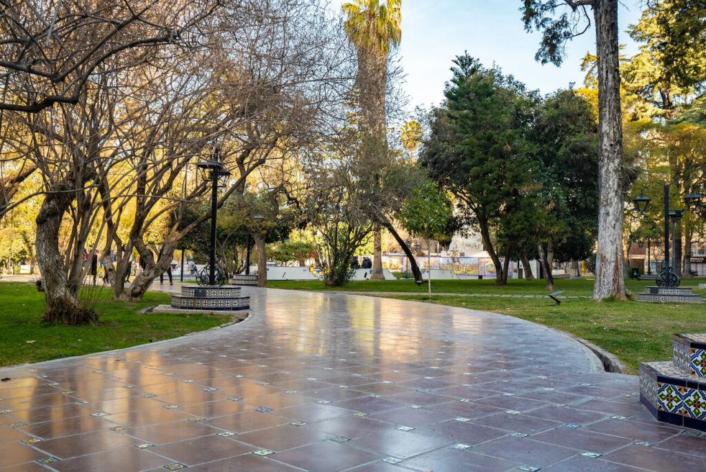 Curved tiled pathway winding through a leafy city park with mature trees, green lawns, decorative tiled benches and lampposts, lit by soft afternoon sunlight and creating a calm urban atmosphere. A helpful scene to consider when deciding where to stay in Mendoza.