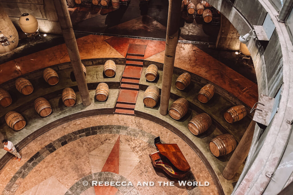Circular underground barrel cellar with rows of wooden wine barrels arranged around tiered levels, viewed from above with a grand piano positioned in the centre of the space during a visit to one of the Mendoza wineries.