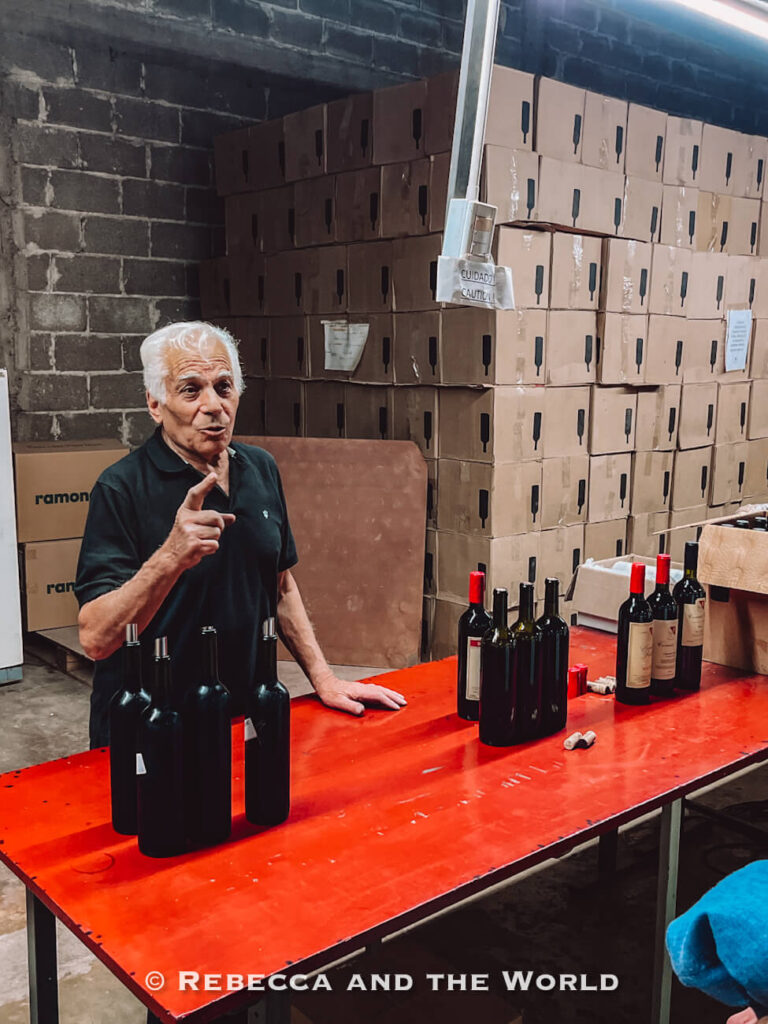 Winemaker Carmelo Patti standing beside a bottling table inside a working winery warehouse, with uncapped wine bottles lined up in front of stacked cardboard boxes during a behind-the-scenes visit to a Mendoza winery.