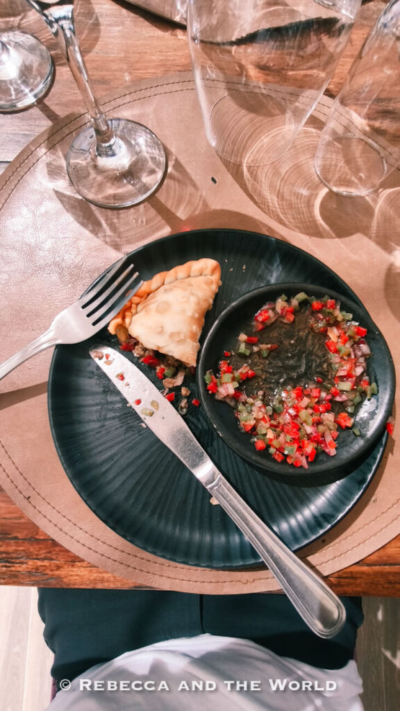 Top-down view of a black plate holding a partially eaten empanada beside a small bowl of chopped red and green salsa, with cutlery, wine glasses, and a wooden table setting around it. A food pairing often enjoyed during Mendoza wine tasting.