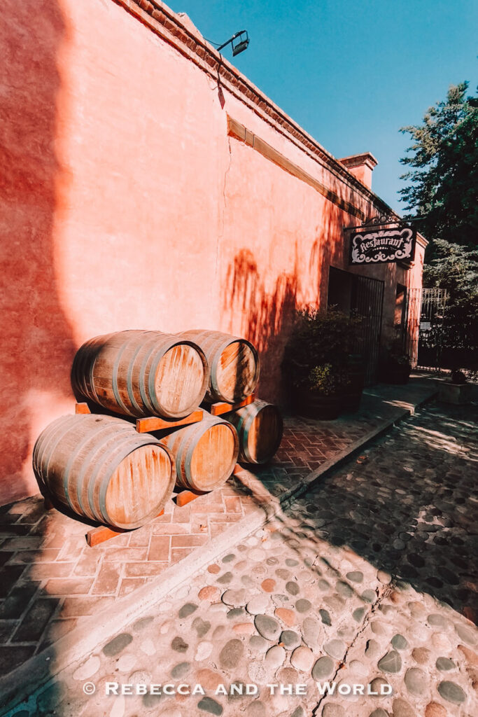 Stacked wooden wine barrels placed outside a rustic pink stucco building with a hanging restaurant sign, cobblestone pathway, and dappled sunlight casting shadows across the scene. A classic setting found at many wineries in Mendoza.