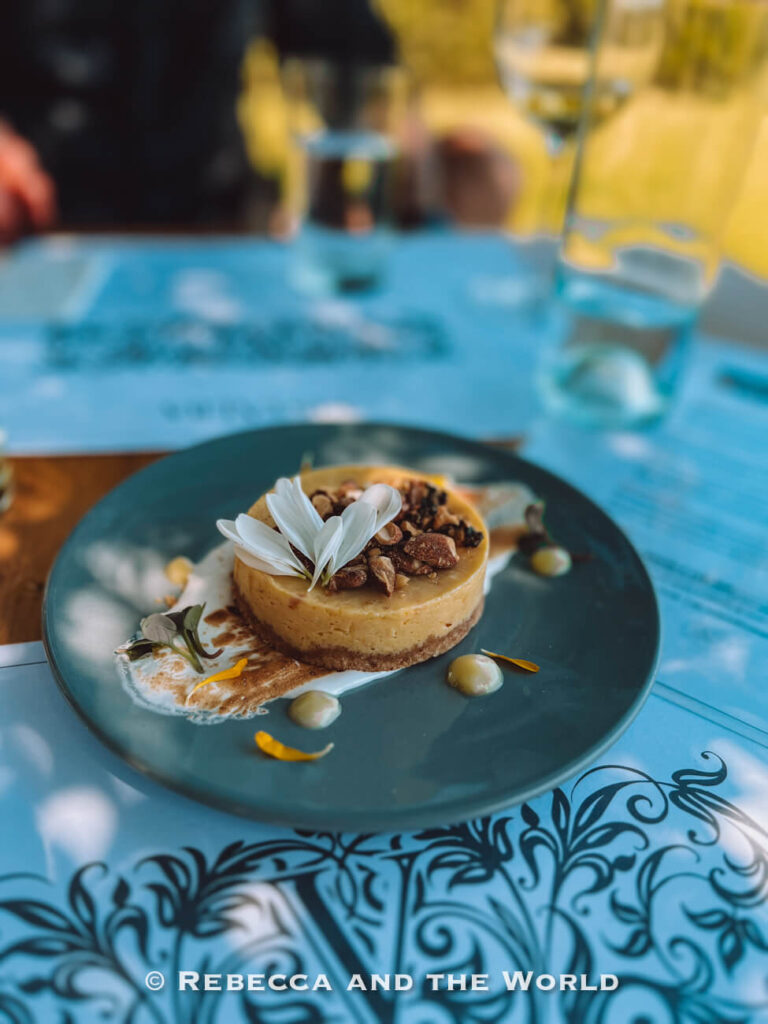 **Close-up of a plated key lime pie served outdoors on a blue patterned table, topped with edible white flower petals and chopped nuts, with decorative sauce and blurred glasses and diners in the background. A beautifully presented dessert to enjoy during Mendoza wine tasting.**