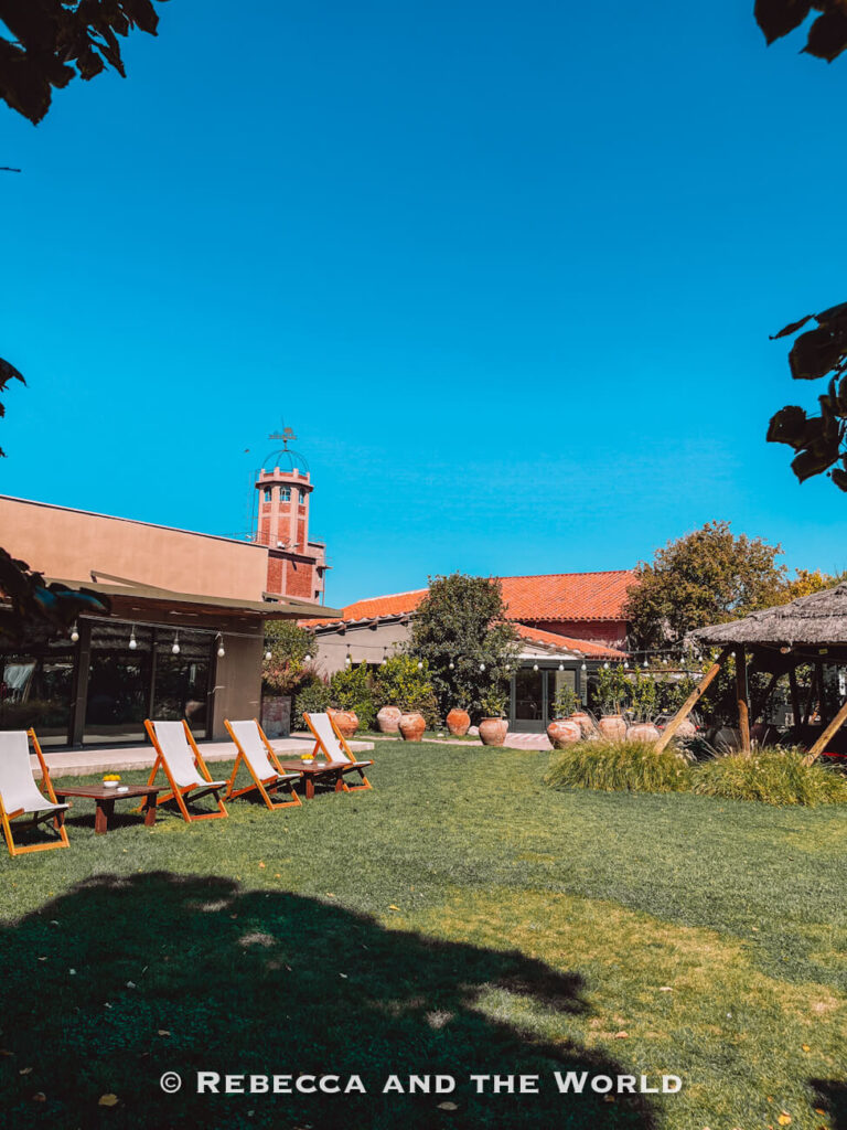 Wide view of the lawn and outdoor seating area at Bodega Kaiken in Luján de Cuyo, with wooden sun loungers arranged on the grass, terracotta pots, low winery buildings with tiled roofs, and the winery tower rising against a clear blue sky. A relaxing stop to include when visiting Mendoza.