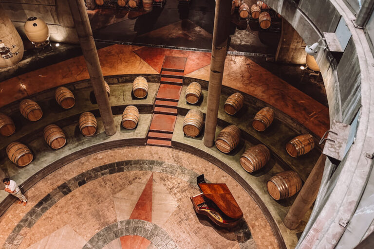 Circular underground barrel cellar with rows of wooden wine barrels arranged around tiered levels, viewed from above with a grand piano positioned in the centre of the space during a visit to one of the Mendoza wineries.