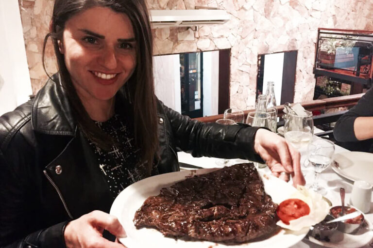 A smiling woman - the author of this article - holding a large plate with a traditional Argentine steak, called an asado, accompanied by a simple garnish of a tomato slice.