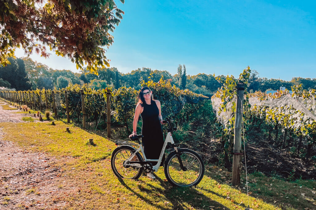 A woman (the author of this blog) standing with a bicycle between vineyard rows in Mendoza, Argentina, on a sunny day with trees and green grass surrounding the path.