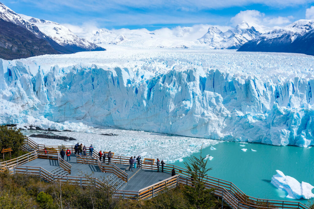 Visitors stand on wooden viewing platforms overlooking the towering blue ice wall of Perito Moreno Glacier in Los Glaciares National Park, Argentina, with turquoise water, snow-covered mountains, and a partly cloudy sky in the background. This is a great stop on when spending 1 week in Argentina.