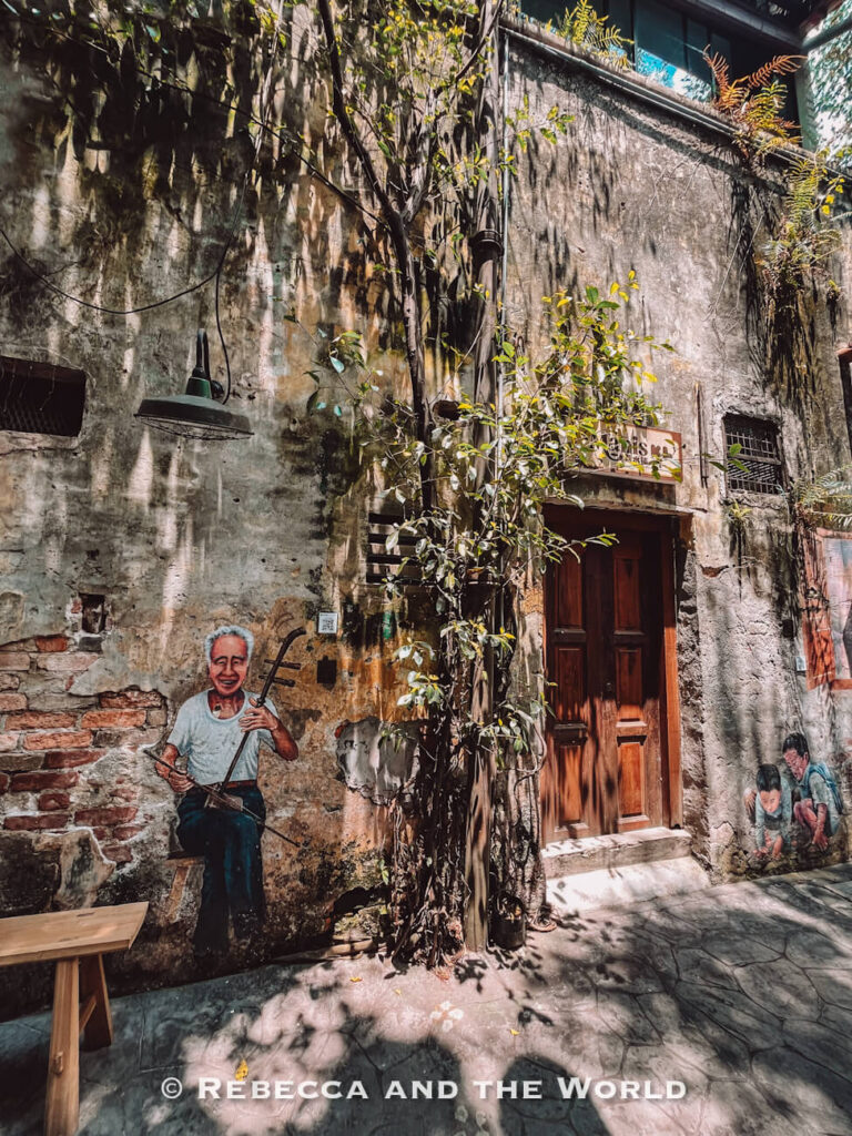 A narrow heritage alley in Kuala Lumpur with weathered concrete and exposed brick walls covered in creeping plants and painted street art. A mural of an elderly man playing a traditional bowed string instrument appears beside a wooden doorway, while another mural of two people is painted near the ground on the opposite side.