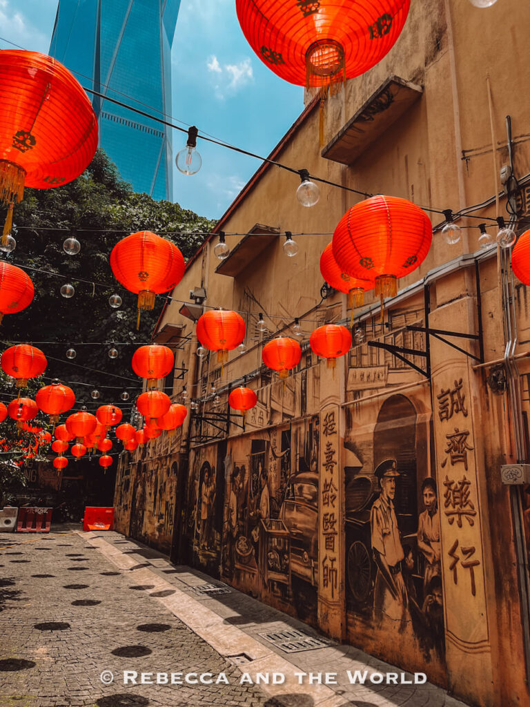 Dozens of bright red Chinese lanterns and strings of clear bulbs hang above a narrow Kuala Lumpur alley lined with sepia-toned murals. The artwork depicts historic street scenes, people, old vehicles and Chinese characters, with a modern glass skyscraper visible beyond the older buildings&mdash;a vivid contrast to include on a Kuala Lumpur itinerary.