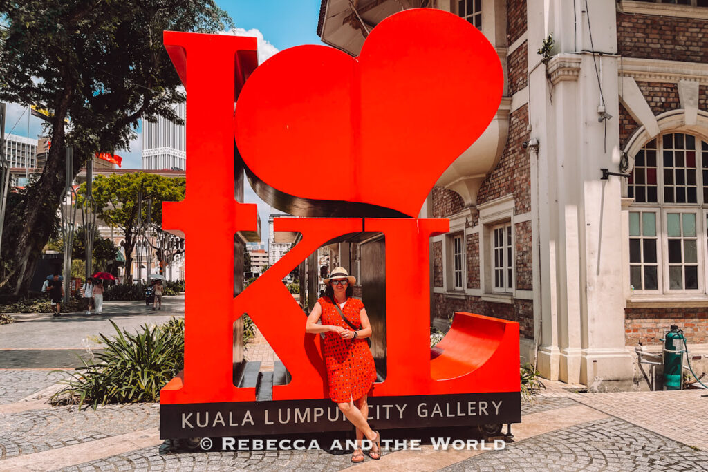 A woman (the author of this article) in a red dress and sun hat stands beneath the giant red &ldquo;I &hearts; KL&rdquo; sculpture outside Kuala Lumpur City Gallery. The bold landmark sits beside a historic brick-and-white building on a sunny day and is a popular photo stop for visitors spending 2 days in Kuala Lumpur.
