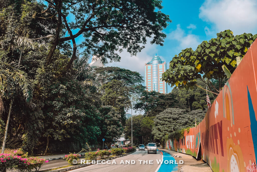 A quiet, tree-lined road in central Kuala Lumpur, Malaysia, with tropical greenery, flowering plants, a bright blue cycle lane and a colorful mural-covered wall. Modern high-rise buildings rise above the dense trees under a vivid blue sky, showing the mix of nature and city architecture visitors can experience during 2 days in Kuala Lumpur.