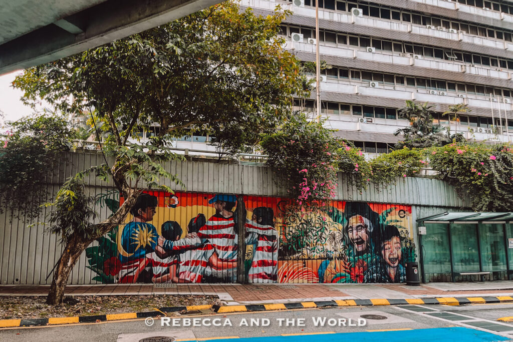 Colorful street art painted across a long roadside wall in Kuala Lumpur, depicting people in Malaysian-themed clothing alongside smiling portraits and tropical plants. Pink flowering vines and leafy trees grow above the mural, with a large city building in the background, making street art a colorful addition to a Kuala Lumpur itinerary.