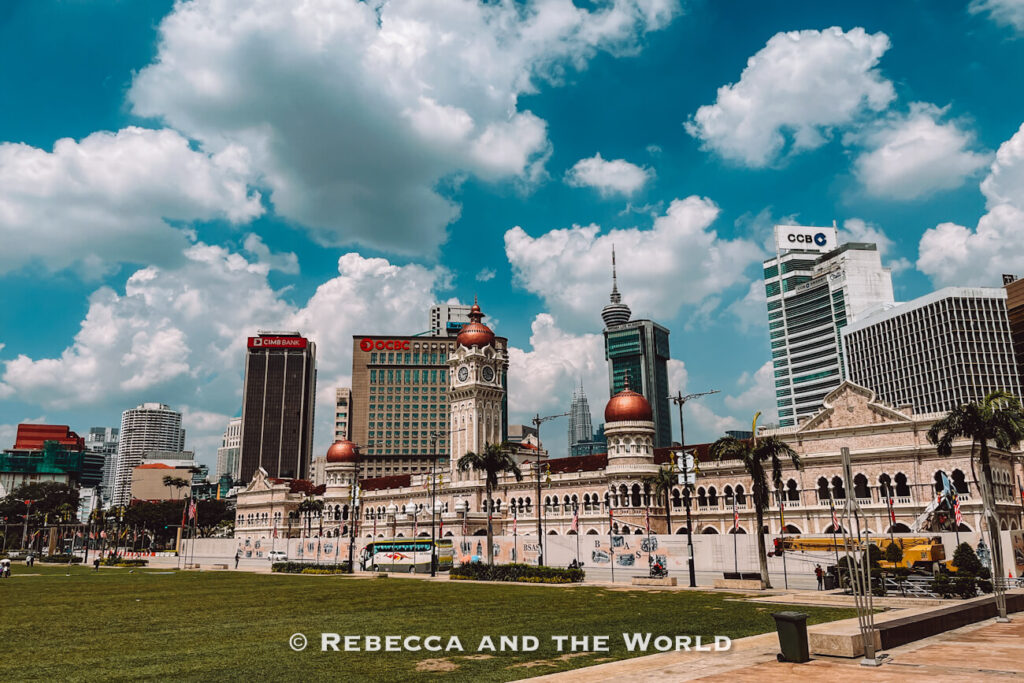 Daytime view across a green lawn toward Kuala Lumpur&rsquo;s historic Sultan Abdul Samad Building, with its arched fa&ccedil;ade, clock tower and copper-colored domes. Modern skyscrapers rise behind the heritage architecture beneath dramatic white clouds, highlighting the contrast between old and new among the things to do in Kuala Lumpur.
