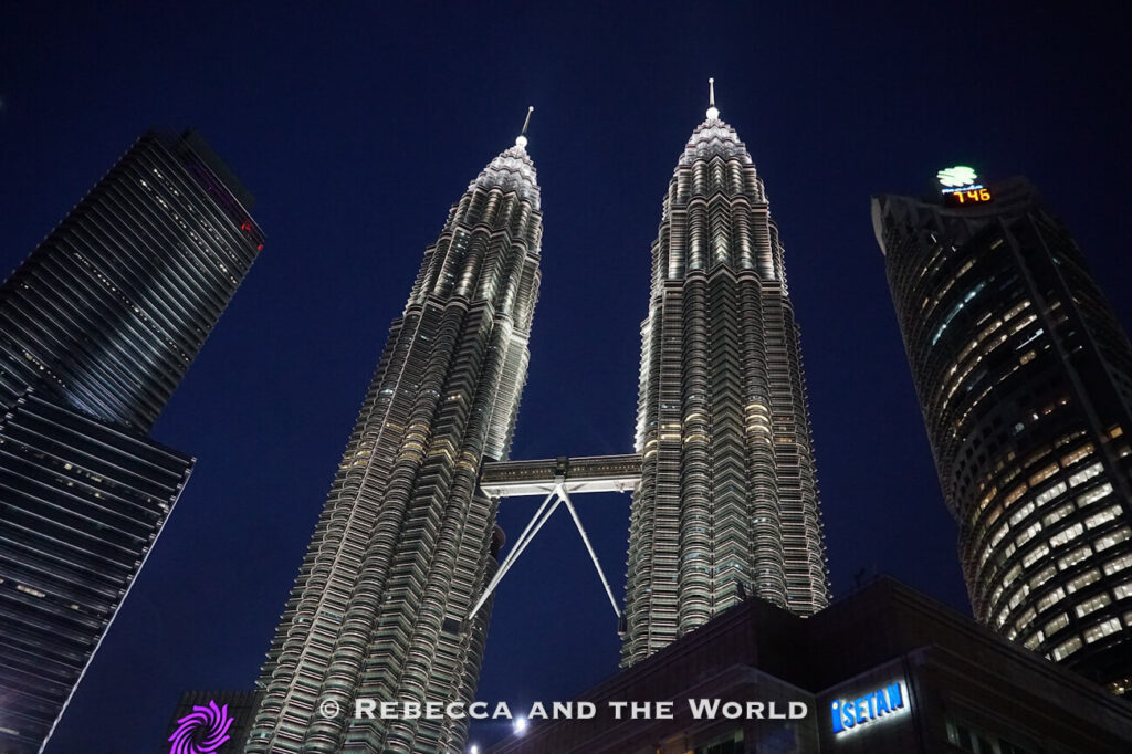 Dramatic low-angle nighttime view of the illuminated Petronas Twin Towers in Kuala Lumpur, their silver fa&ccedil;ades glowing against a deep navy sky. The skybridge connects the two towers above surrounding high-rises, creating one of the city&rsquo;s most recognizable sights for a Kuala Lumpur itinerary.