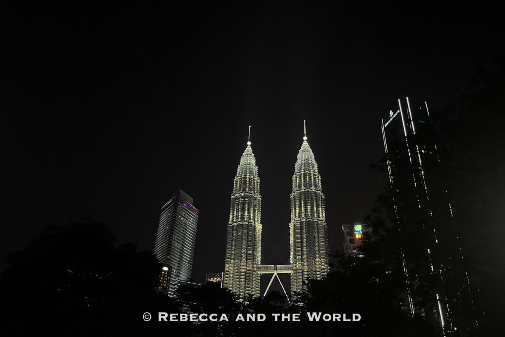 The illuminated Petronas Twin Towers rise above dark trees and surrounding skyscrapers in the Kuala Lumpur skyline at night. Their bright twin spires and connecting skybridge stand out against the black sky, making the landmark a memorable evening sight during 2 days in Kuala Lumpur.