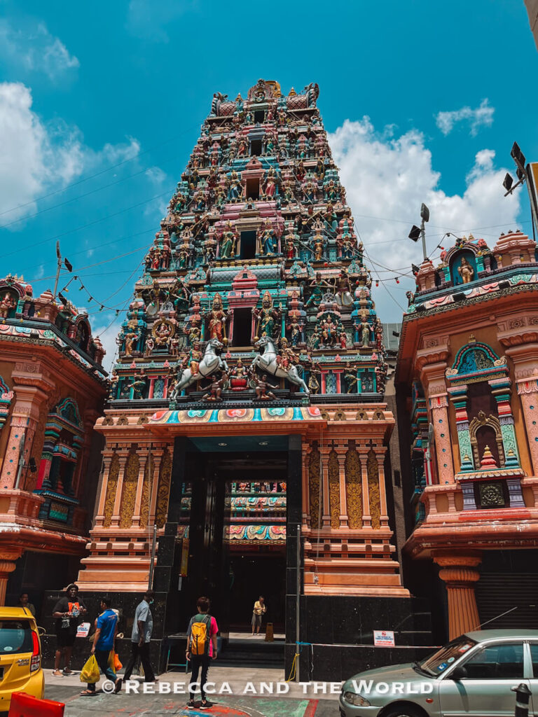 Elaborately decorated entrance tower of a Hindu temple in Kuala Lumpur, covered from top to bottom with colorful sculptures of Hindu deities, figures and animals. Pink, turquoise, gold and green architectural details stand out against a bright blue sky, while several visitors walk near the entrance below&mdash;one of the striking cultural things to do in Kuala Lumpur.