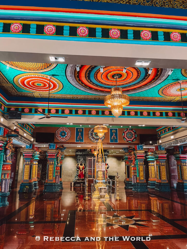 Ornate interior of a Hindu temple in Kuala Lumpur, with brightly painted turquoise, red, gold and green columns, decorative religious sculptures and elaborate circular ceiling designs. Golden chandeliers hang above a polished dark floor that reflects the richly decorated interior.