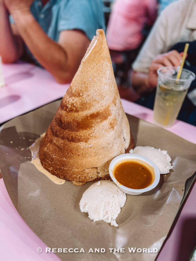 Tall cone-shaped Indian dosa served on brown paper with coconut chutney and sambar at a casual eatery in Little India, Singapore.