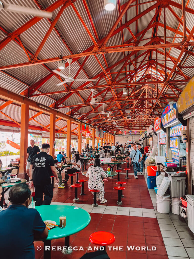 Diners eating at green tables inside a busy Singapore hawker centre beneath a high roof supported by bright orange steel beams. This is Maxwell Food Centre, one of the best hawker centres in Singapore.