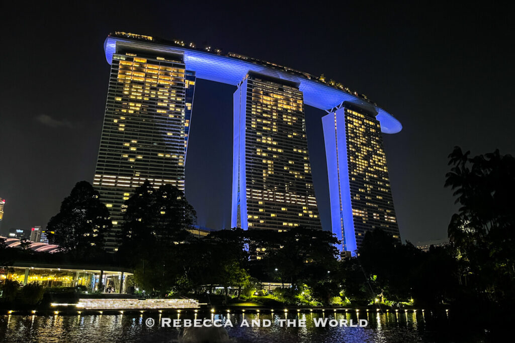 Marina Bay Sands illuminated in blue and gold at night, viewed across the water and gardens from Gardens by the Bay.