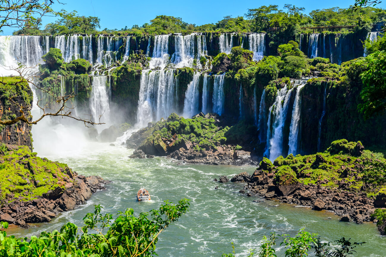 Wide view of Iguazu Falls on the Argentina–Brazil border, with multiple waterfalls cascading over lush green cliffs into the river below and a sightseeing boat approaching the falls. Iguazu Falls is a must-visit on your 4 weeks in Argentina itinerary.