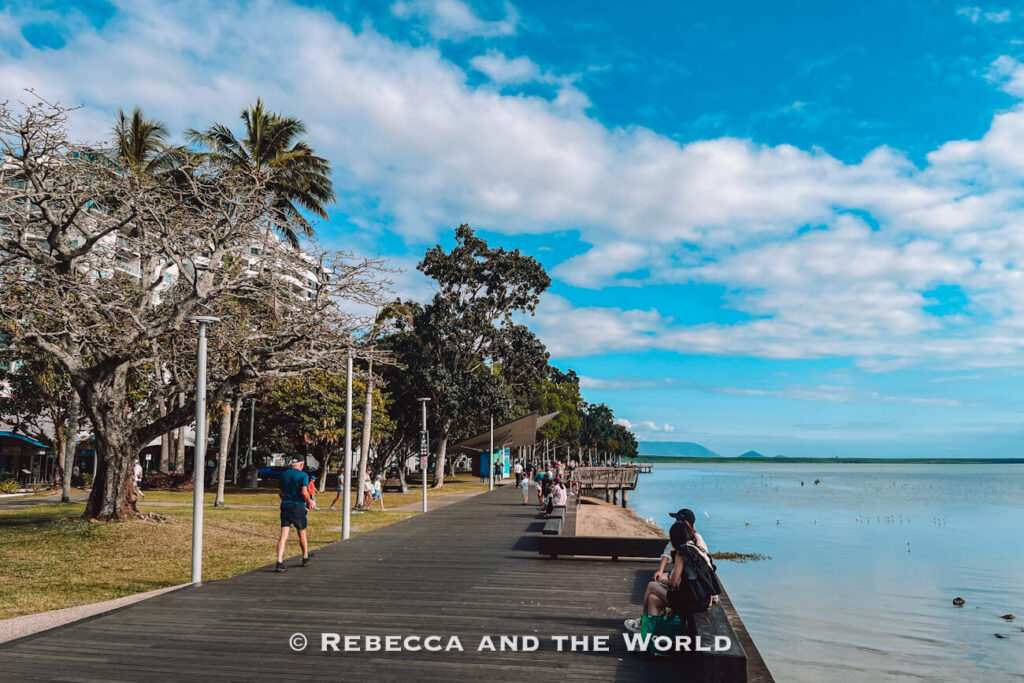 The Cairns Esplanade boardwalk on a sunny day, with people walking and sitting on benches along the timber path, tropical trees and palms on the left, and the calm waters of Trinity Bay stretching out to distant hills on the right.
