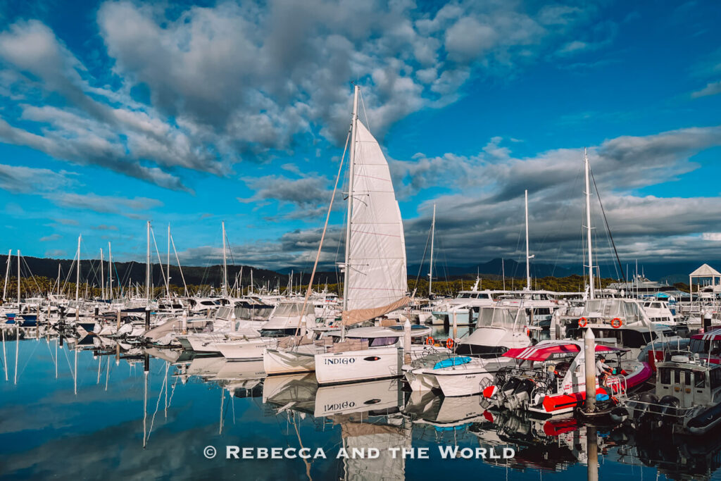 Sailing catamaran named Indigo with its white sail raised, moored among other boats at Crystalbrook Marina under a partly cloudy sky, the departure point for Great Barrier Reef boat trips