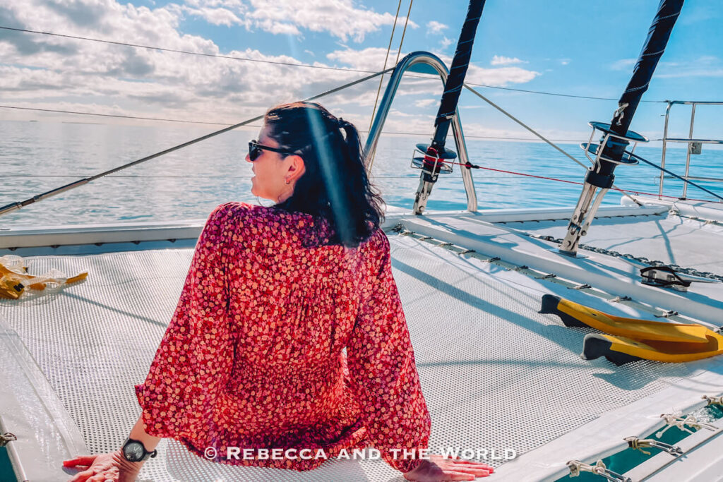 Passenger (the author of this article) sitting on the trampoline netting at the front of the Sailaway catamaran with snorkelling flippers nearby, sailing out from Port Douglas towards the Great Barrier Reef