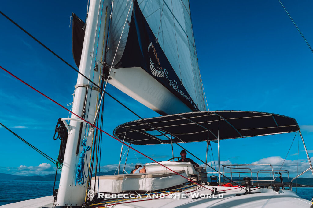 The Sailaway catamaran's captain at the helm under the shaded cockpit roof, with the branded sail raised against a clear blue sky