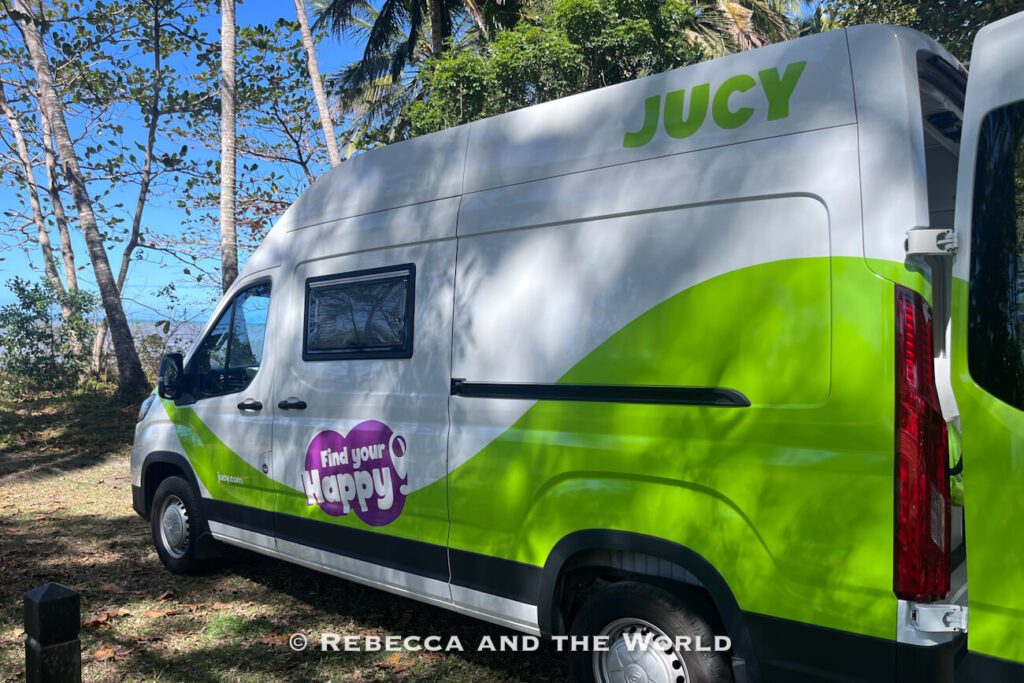 Side view of a green and white Jucy campervan with its "Find Your Happy" branding, parked among palm trees on a Tropical North Queensland road trip