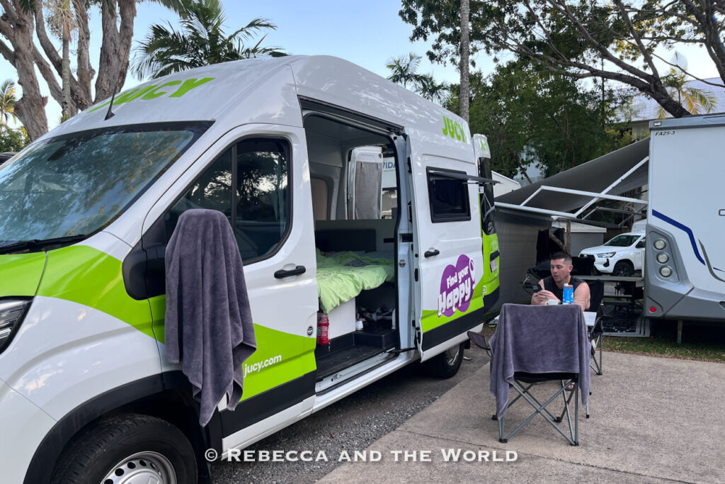Jucy campervan with its side door open showing the made-up bed, parked at a caravan park in Port Douglas with camping chairs and a table set up alongside