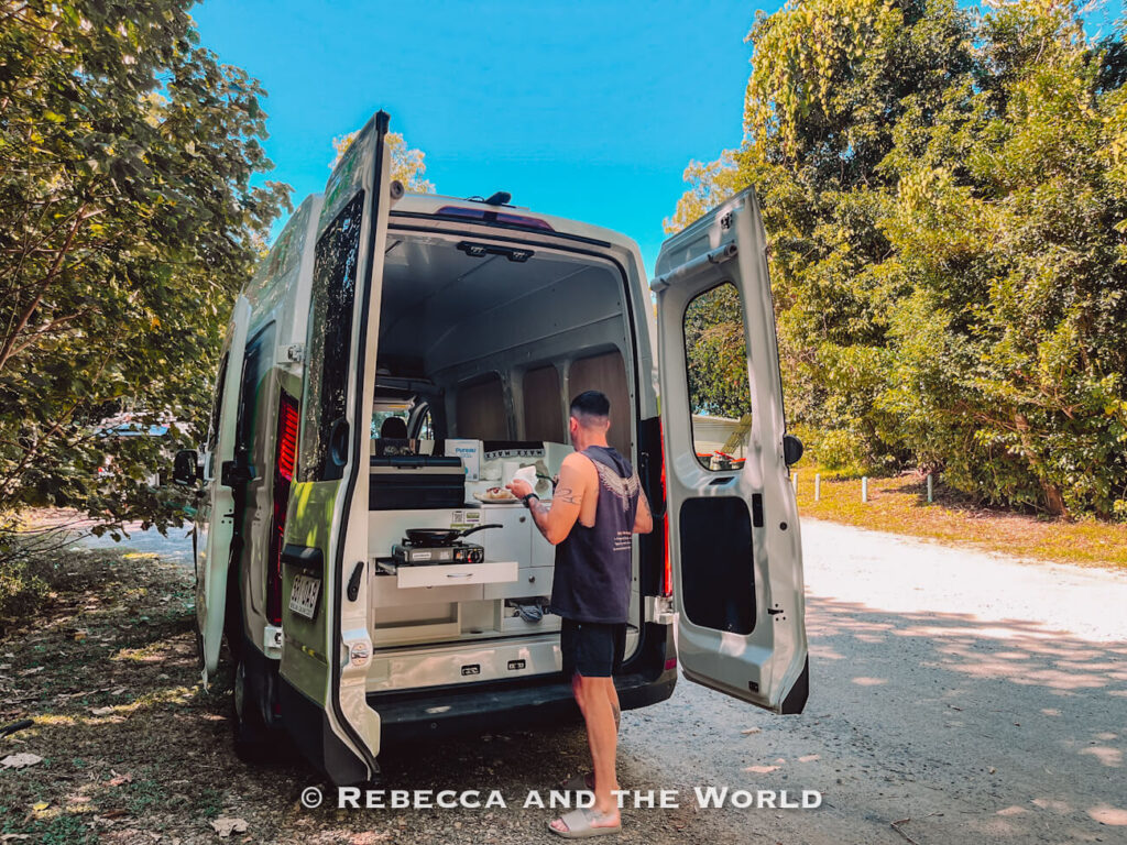 Man (the author’s husband) preparing lunch at the pull-out kitchen in the back of a Jucy campervan, with the gas stove and frying pan visible through the open rear doors
