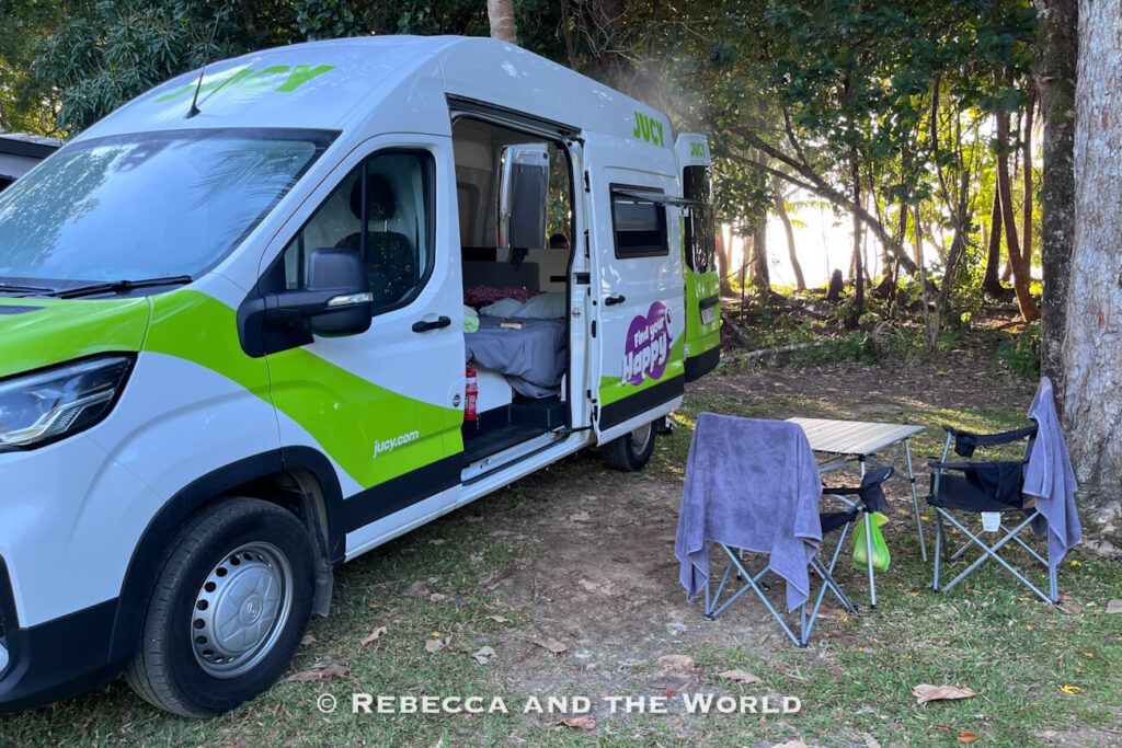 Jucy campervan parked at a Cape Tribulation campsite with its side door open, camping chairs and a table set up beside it in the dappled light