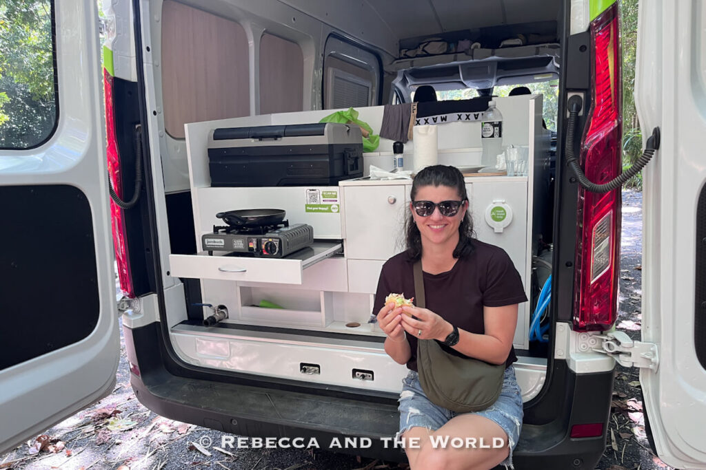Woman (the author of this article) sitting at the rear of a Jucy campervan eating lunch, with the fitted kitchen and fridge visible behind her
