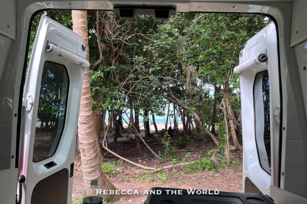 View through the open rear doors of a Jucy campervan out to rainforest trees with a glimpse of the beach beyond, parked near Cape Tribulation