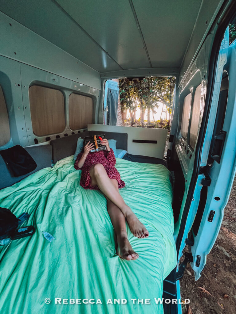 Woman (the author of this article) relaxing and reading on the made-up double bed inside a Jucy campervan, with the side door open to the rainforest outside