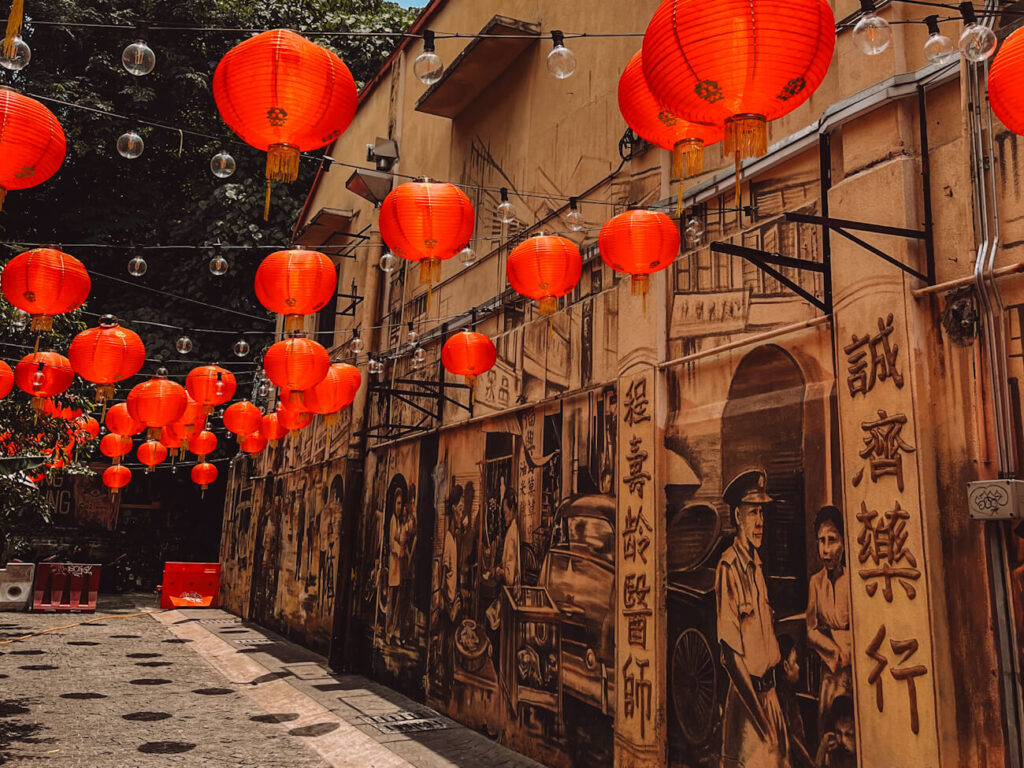 Dozens of bright red Chinese lanterns and strings of clear bulbs hang above a narrow Kuala Lumpur alley lined with sepia-toned murals. The artwork depicts historic street scenes, people, old vehicles and Chinese characters.
