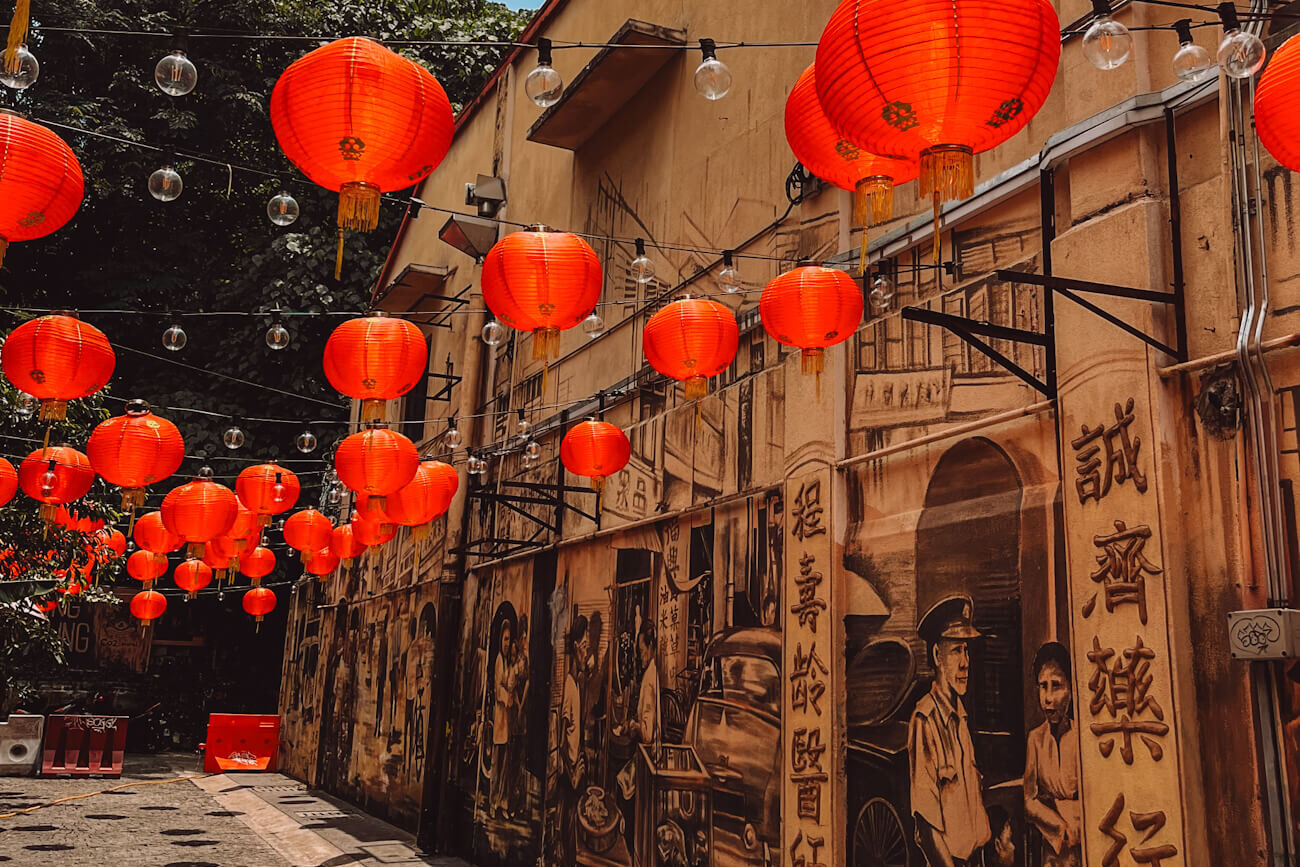 Dozens of bright red Chinese lanterns and strings of clear bulbs hang above a narrow Kuala Lumpur alley lined with sepia-toned murals. The artwork depicts historic street scenes, people, old vehicles and Chinese characters.
