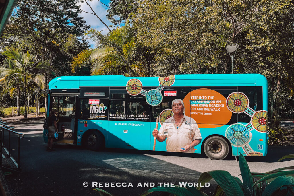 The bright turquoise Mossman Gorge shuttle bus, painted with Indigenous artwork and a "100% electric" sign, parked at the Mossman Gorge Centre with a passenger boarding