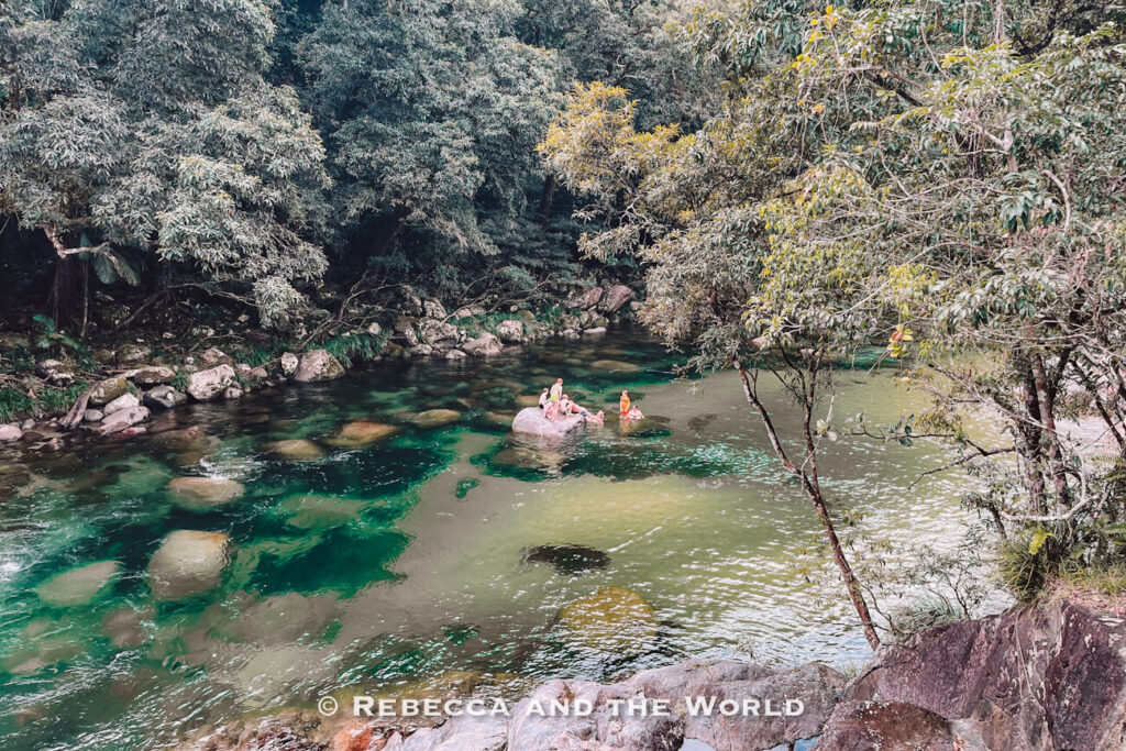 Swimmers sitting on a boulder in the clear green water of the Mossman River, the main Mossman Gorge swimming spot, surrounded by rainforest